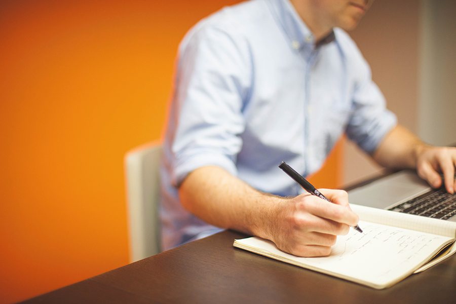 Man on his laptop with a pen and notebook to his right as he is writing information down